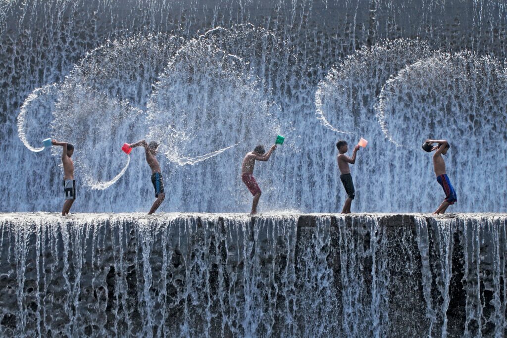 Five boys using buckets to splash water at each other during Songkran, set in a waterfall.