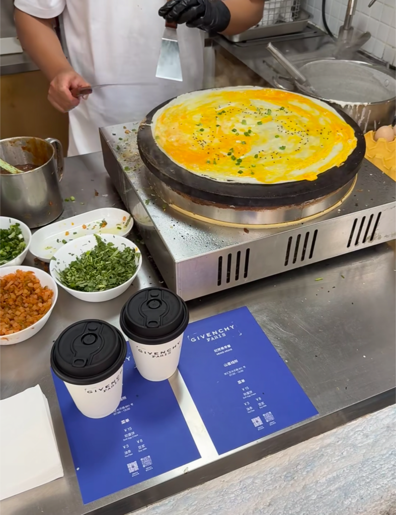 Givenchy's 2026 Shanghai pop-up: 纪梵希早餐 (Givenchy Breakfast) - 山喜线所 (Shanxi Noodle Shop) shows a man making a 软蛋饼 (Soft Egg Crepe) with 豆浆 (Soy Milk) on the side